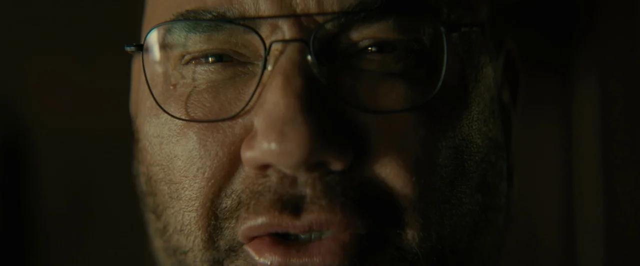 Knock at the Cabin (2023) movie still: closeup — A dimly lit, shadow-filled environment frames a man's face in tight focus. He holds a direct gaze…