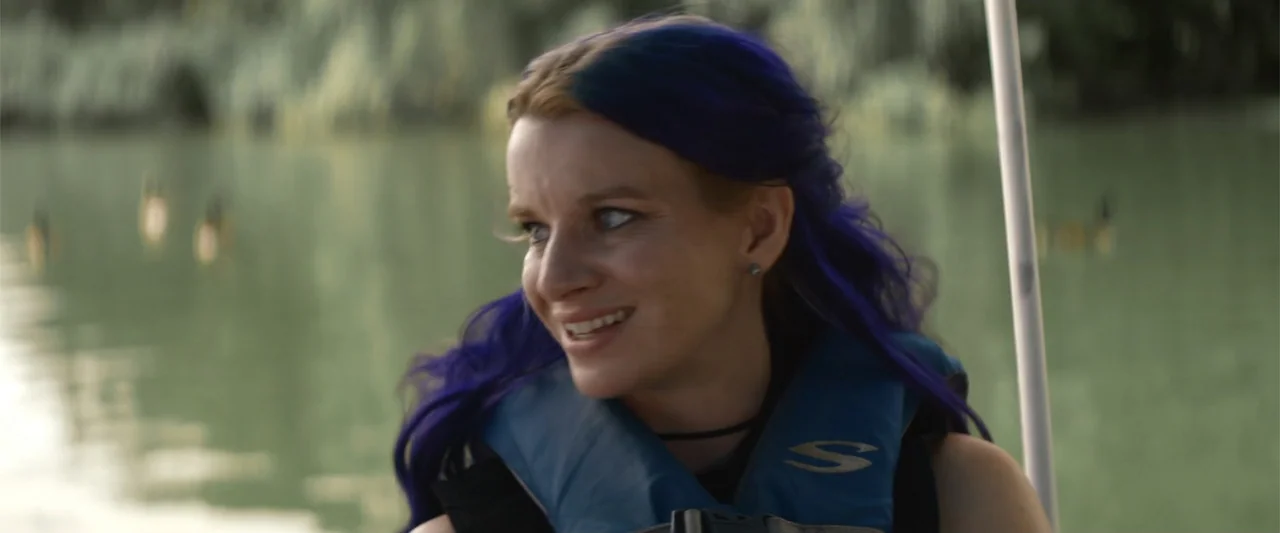 Mennonite's Bite (2023) movie still: medium — A woman sits in a vessel on calm water with blurred trees in background. The subject wears a blue life…