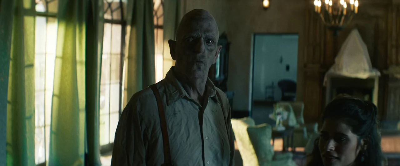 The Mad Hatter (2021) movie still: medium — An aged man with a pale, bald head stands in a dimly lit, vintage parlor. He looks forward while a woman…
