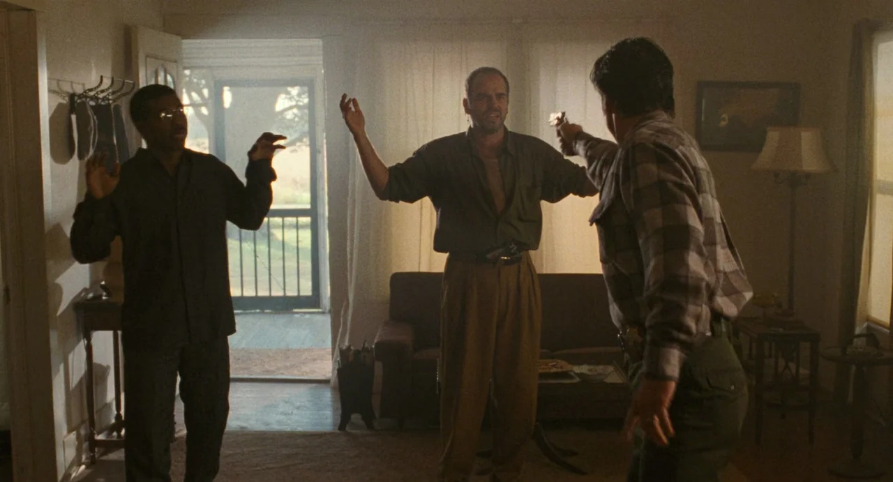 One False Move (1991) movie still: fullbody — A cluttered interior space with a couch, a lamp, and a screen door entrance. A man holds a gun on two…