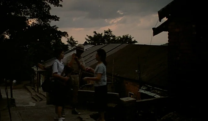 Dust In The Wind (1986) movie still: fullbody — A rural outdoor scene with a dark building and overcast sky at twilight. Three young people gather…