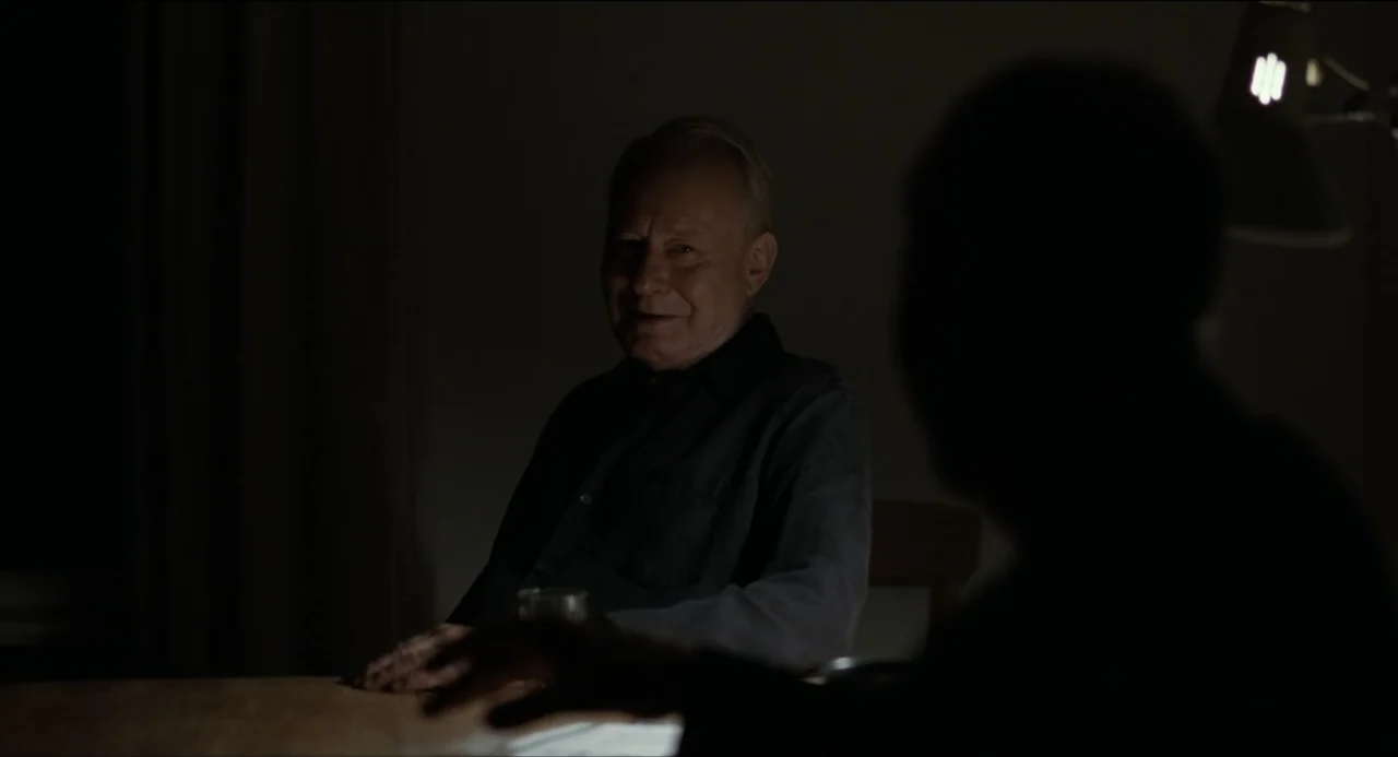 Sentimental Value (2025) movie still: medium — A dimly lit room with an older man sitting at a wooden table. The man faces a blurred silhouette in…