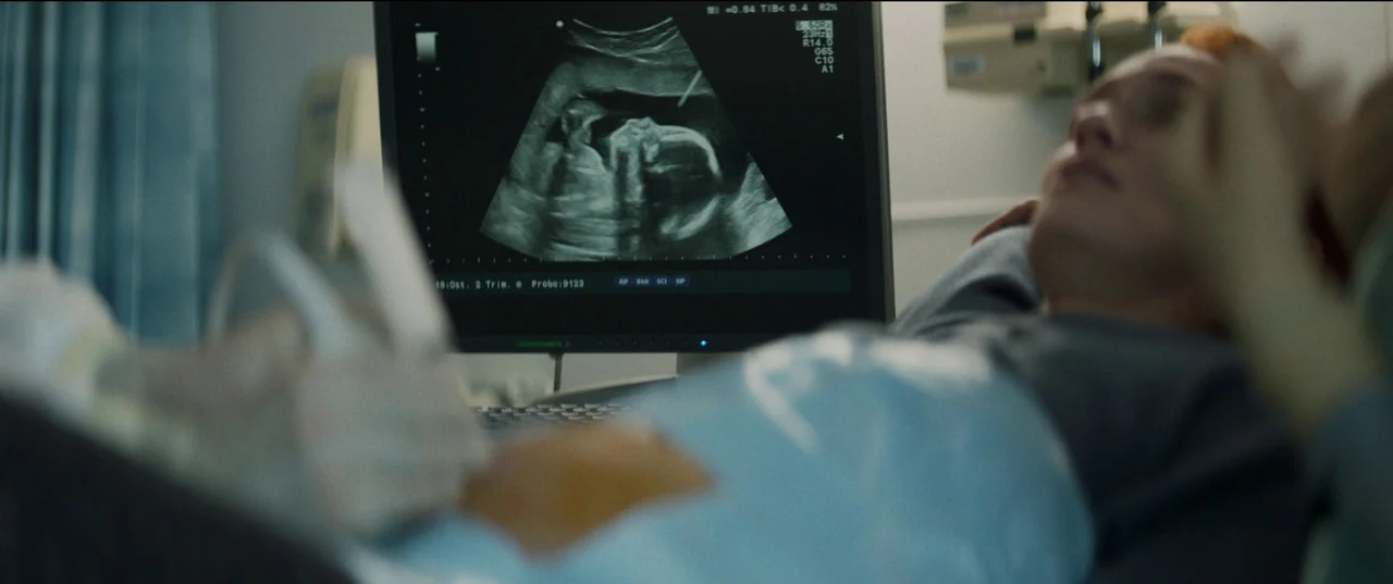 Birth/Rebirth (2023) movie still: medium — A medical exam room contains a large monitor displaying an ultrasound image of a fetus. A person lies on…