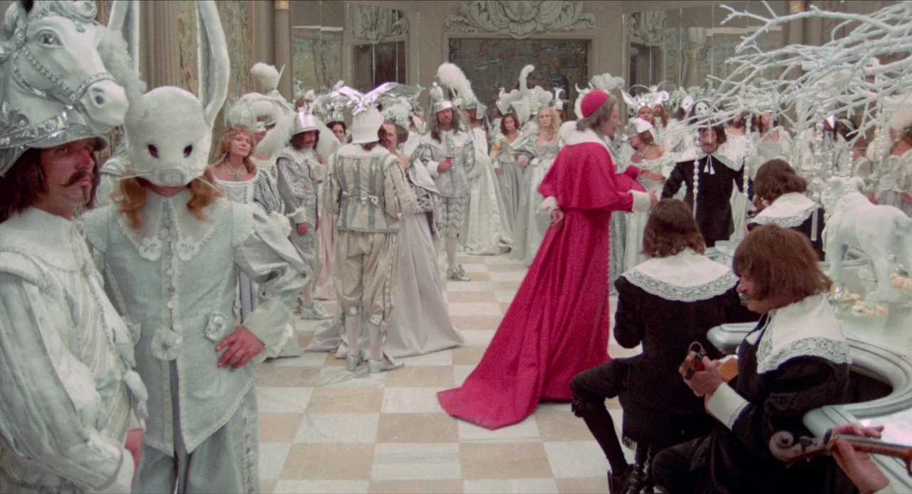 The Three Musketeers (1973) movie still: wide — A crowded ballroom floor features guests in white monochromatic masquerade costumes and animal…