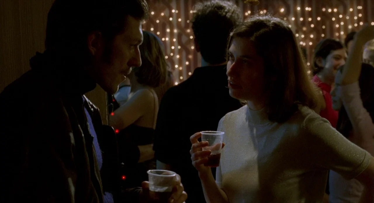 Read My Lips (2001) movie still: medium — A crowded social gathering occurs in a dimly lit room with twinkling fairy lights. A man and a woman stand…