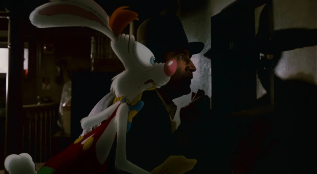 Who Framed Roger Rabbit (1988) movie still: medium — A dimly lit room serves as the setting for an encounter between two figures. A human man in a…
