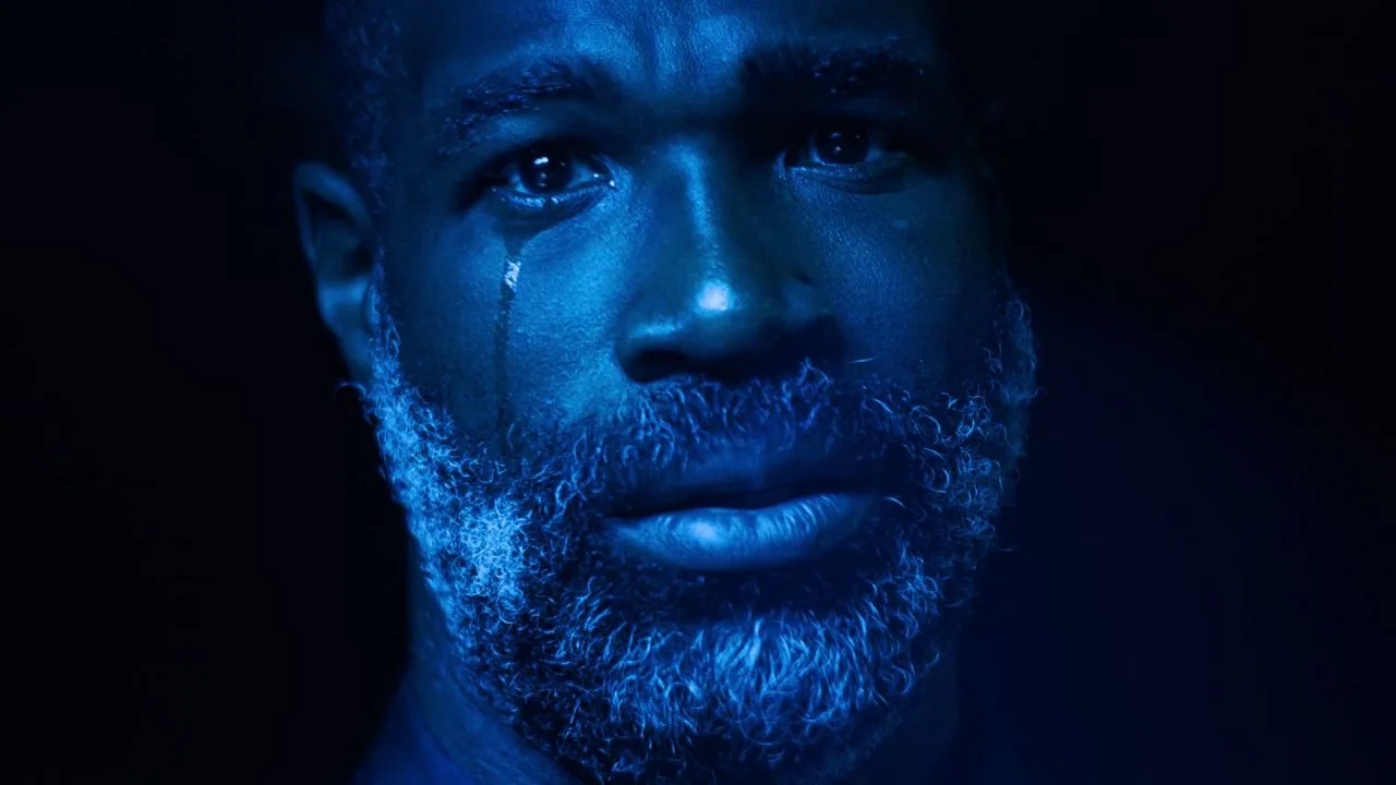 She Dies Tomorrow (2020) movie still: closeup — A man's face is centered against a pitch-black background. A single visible tear tracks down his…
