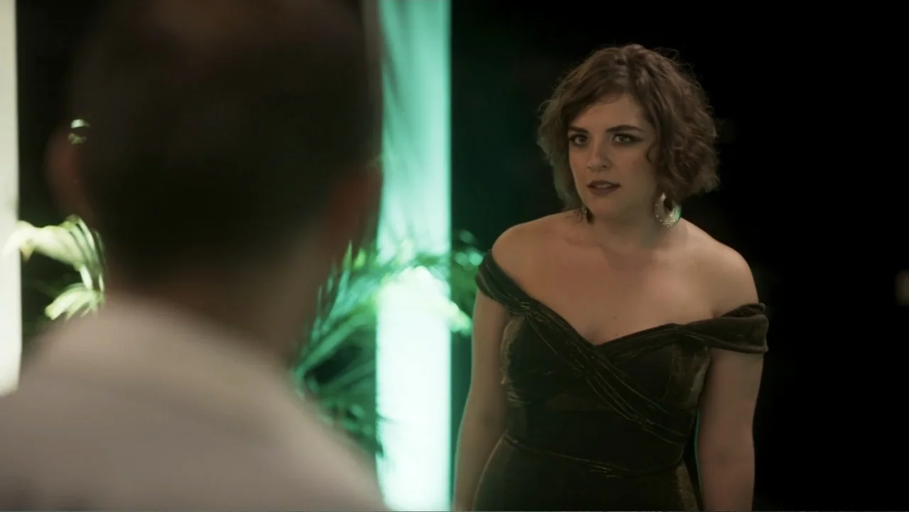 Lecciones para canallas (2022) movie still: medium — A dark event hall features a bright vertical green neon light in the background. A woman in an…