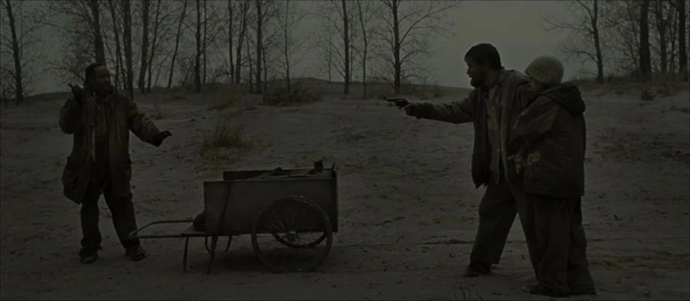 The Road (2009) movie still: wide — A desolate, treeless landscape covered in dry soil under a grey overcast sky. An armed man confronts a stranger…