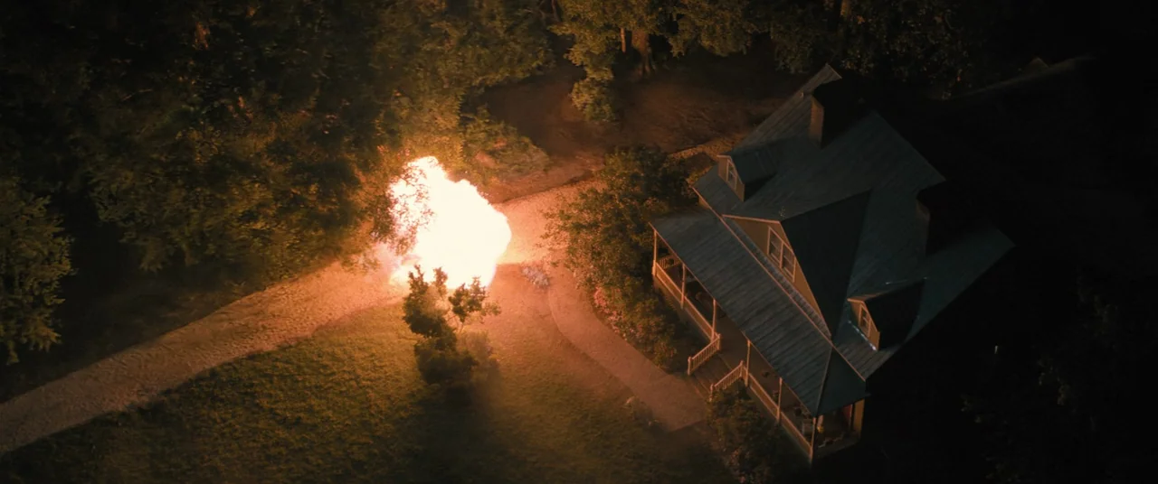 No One Will Save You (2023) movie still: establishing — A two-story house stands on a dark lawn at night near a dense wood. A bright explosion…