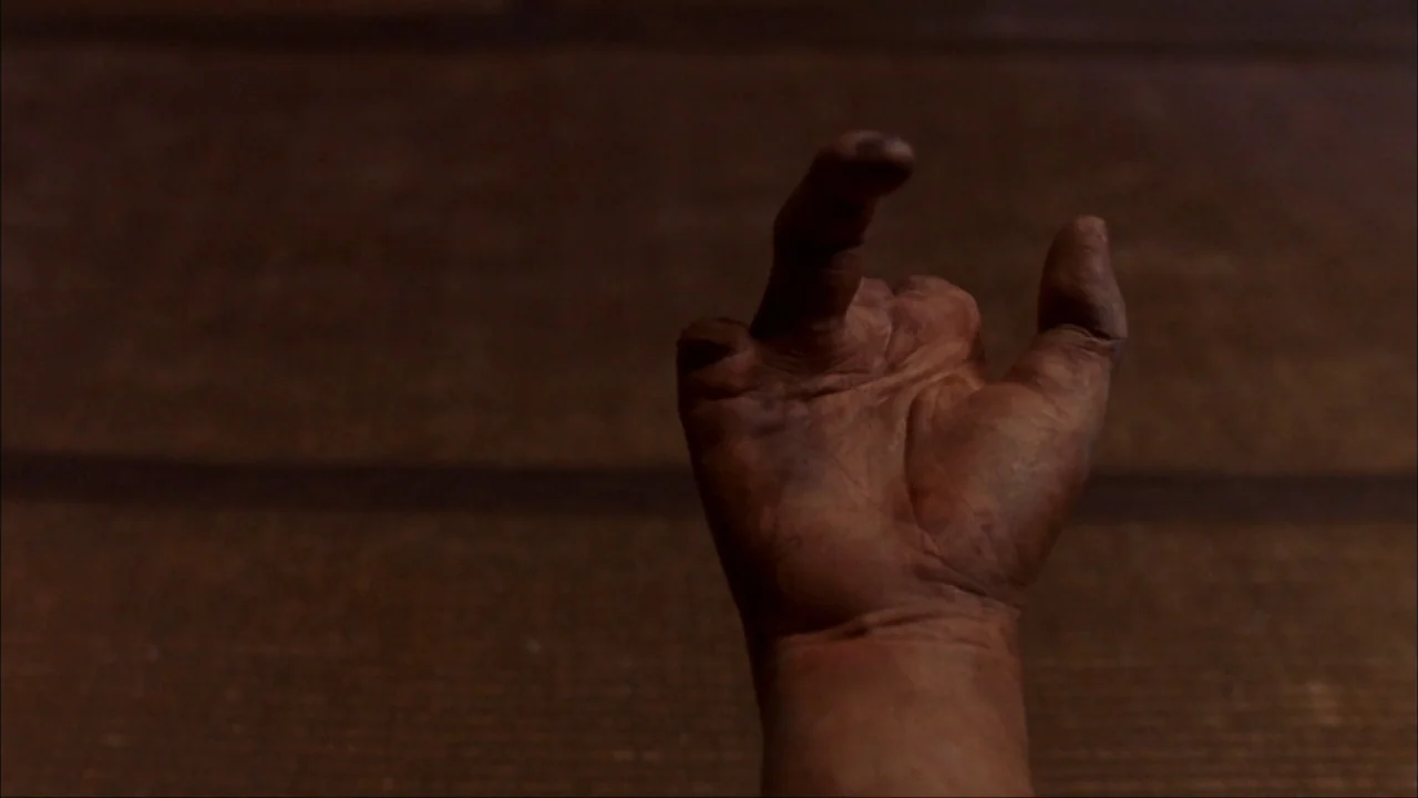 Audition (1999) movie still: closeup — A dark, textured background frames a single mud-covered hand raised into the frame. The fingers are bent in a…