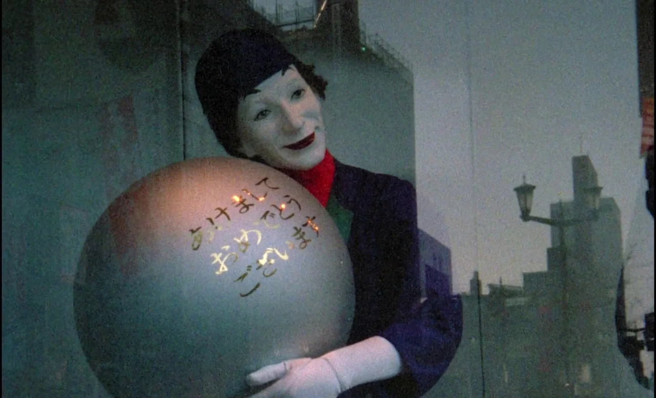 Sans Soleil (1983) movie still: medium — A person with white face makeup stands inside a storefront display holding a sphere. The reflection of city…