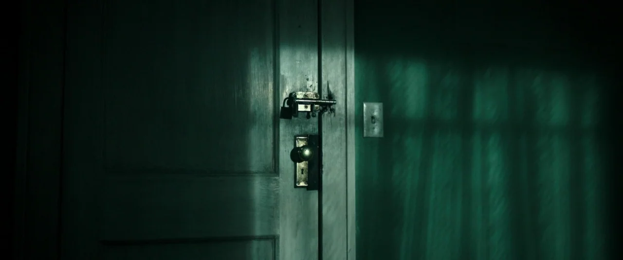 Don’t Breathe (2016) movie still: closeup — A wooden door with a heavy padlock and deadbolt is set into a wall. The scene is illuminated by a…
