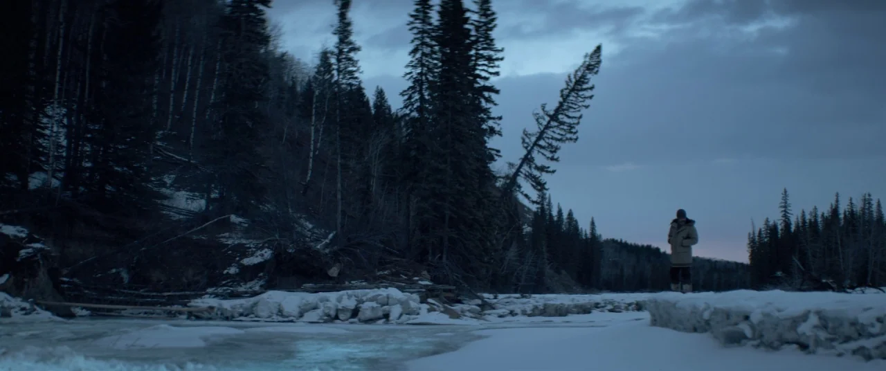 Hold The Dark (2018) movie still: wide — A snowy riverbank borders a dense coniferous forest under a fading twilight sky. A lone man in a parka…