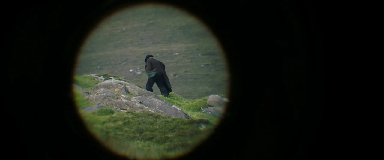 The Banshees of Inisherin (2022) movie still: wide — A grassy, rocky hillside stretches under an overcast sky. A person walks up the slope viewed…