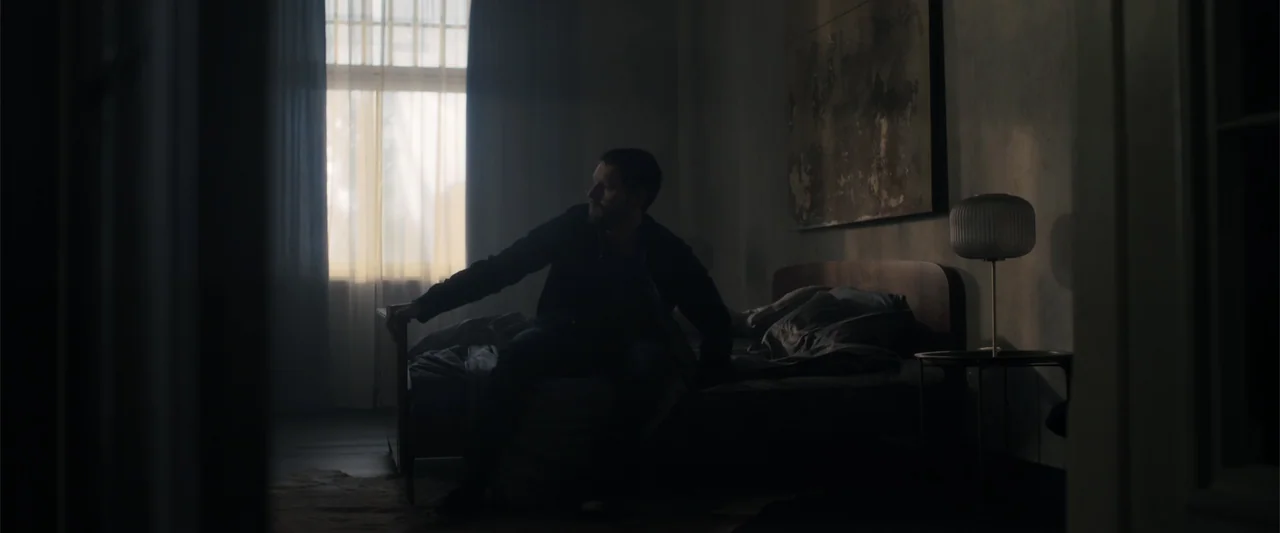 The Good Neighbor (2022) movie still: medium — A dimly lit room contains a bed against a wall with a large abstract painting. A man sits on the edge…