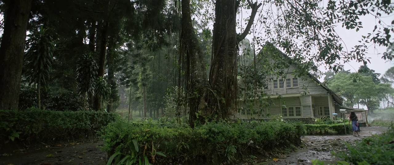Satan’s Slaves (2017) movie still: wide — A wooden building sits nestled among tall trees in a rural, misty environment. A person walks along a…