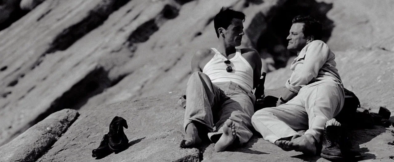 A Single Man (2009) movie still: fullbody — Two men sit together on a large, sunlit rock formation with a steep slope. They are talking to each…