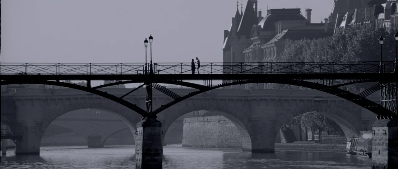 Angel-A (2005) movie still: wide — A wide shot displays the Pont des Arts bridge stretching across the Seine river. Two figures stand as black…