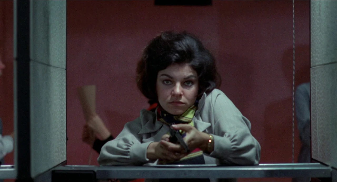 Medium Cool (1969) movie still: medium — A woman stands behind a metallic lane divider inside a dimly lit indoor facility. She holds a compact black…