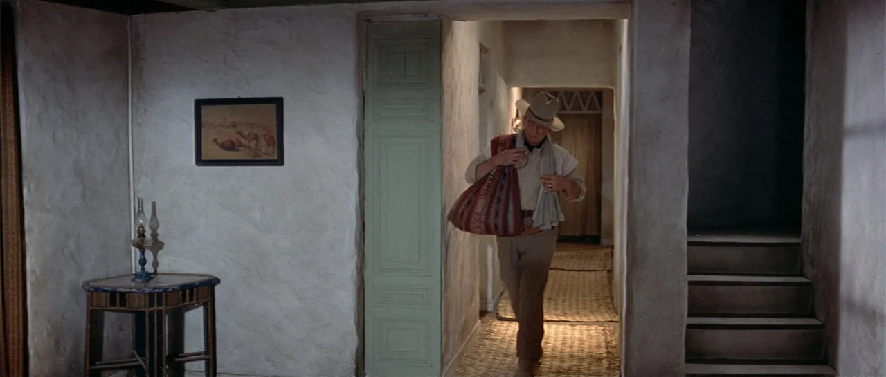 Legend of the Lost (1957) movie still: fullbody — A rustic hallway with white plaster walls and straw floor mats leads toward an interior room. An…