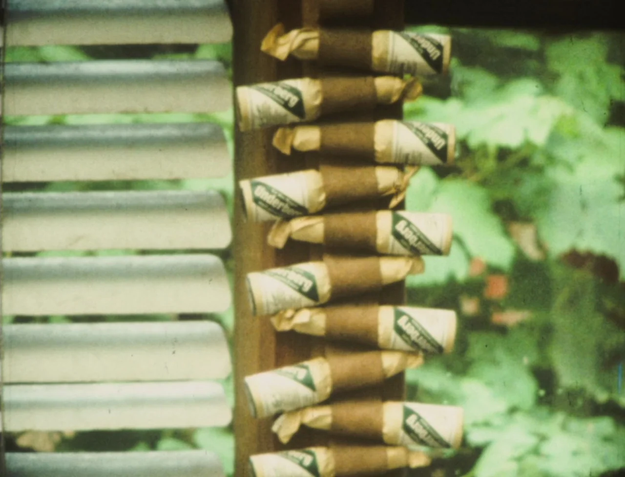 Nekromantik (1988) movie still: closeup — A vertical wooden beam holds a series of small paper-wrapped bottles outdoors. The background consists of…