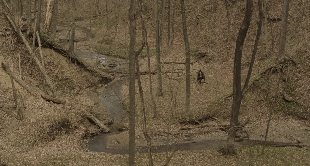 The Alchemist Cookbook (2016) movie still: wide — A deep wooded ravine features a winding streambed surrounded by bare, leafless deciduous trees. A…