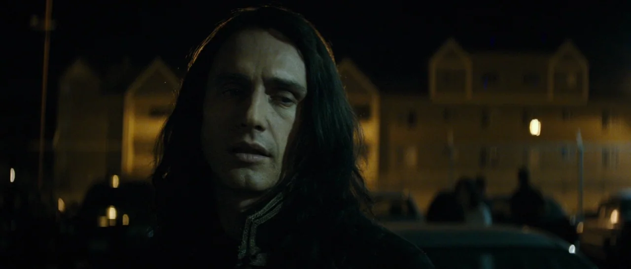 The Disaster Artist (2017) movie still: closeup — A nighttime parking lot exterior with a building silhouette in the distance. A man with long dark…