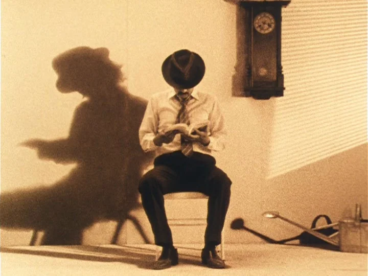 The Woman with Two Heads (1977) movie still: fullbody — A plain room features a wall-mounted clock and a floor-level light source. A man in a hat…