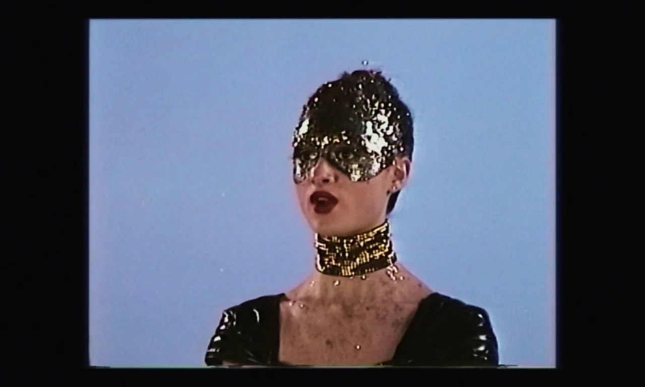 Vox Lux (2018) movie still: closeup — A studio setting with a solid light blue background wall. A woman wears a gold sequined mask and matching gold…
