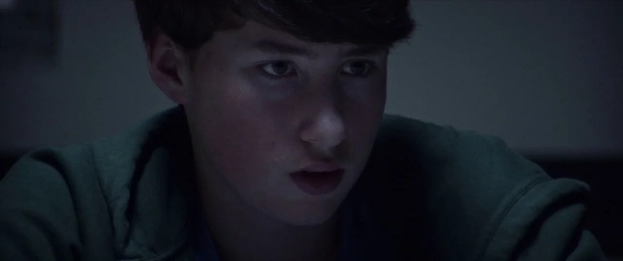 Assassination Nation (2018) movie still: closeup — A dark, dimly lit indoor setting is visible in the frame. A young man looks down at a source of…