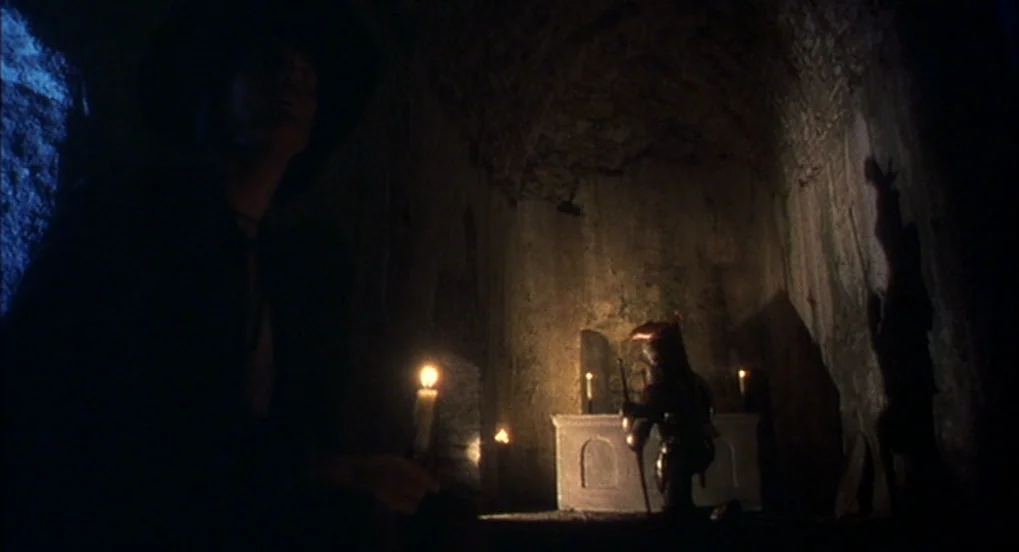 Jabberwocky (1977) movie still: medium — A dark stone crypt features a small altar with lit candles. A cloaked individual observes a knight in plate…