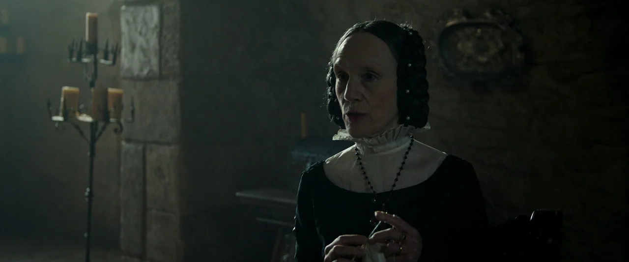 The Last Duel (2021) movie still: medium — A stone-walled interior space with heavy, dimly lit atmosphere suggestive of an 1800s building. An older…
