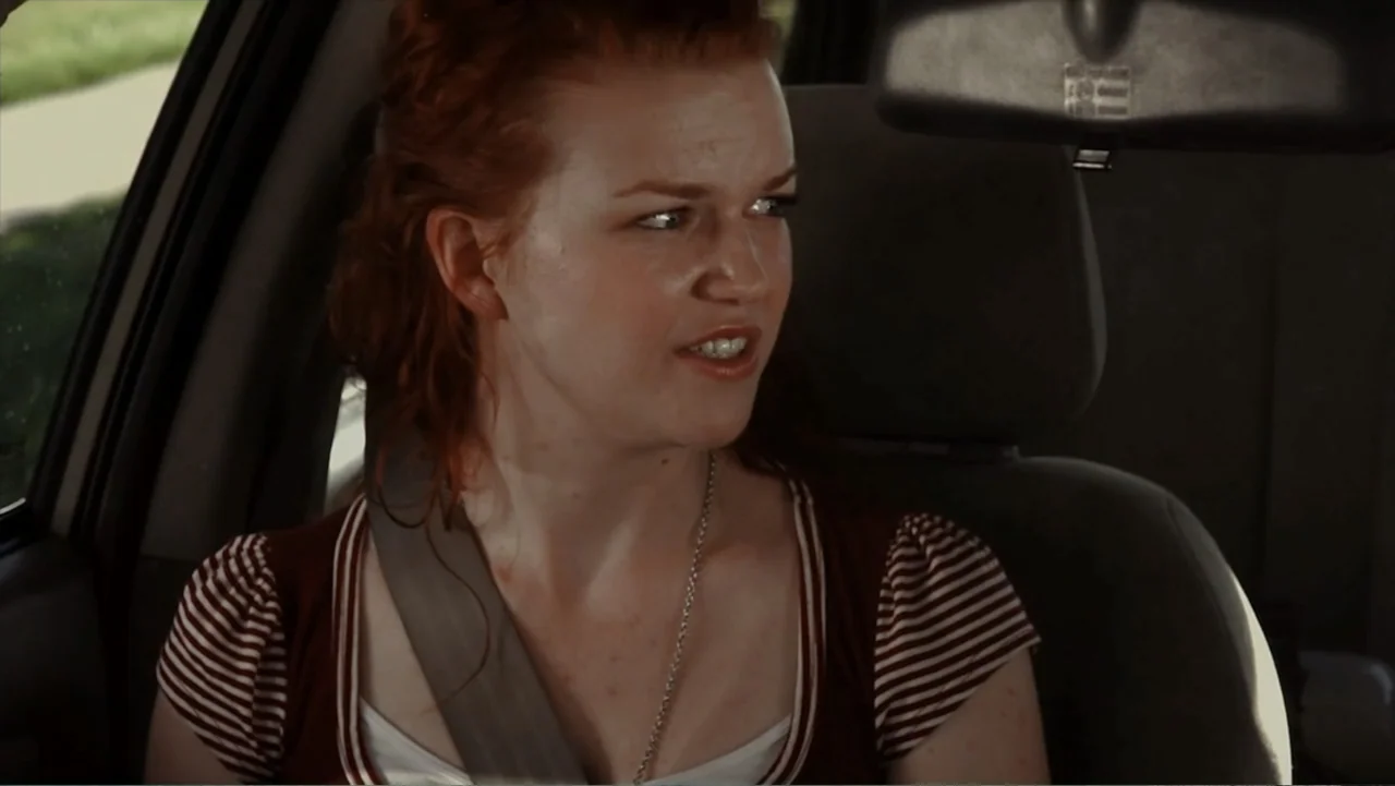 Hello I Love You (2018) movie still: medium — The interior of a car features grey upholstery and a standard rearview mirror. A young woman with red…