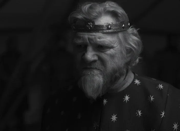 The Tragedy of Macbeth (2021) movie still: closeup — An older man wearing a metal crown stares ahead in a dark setting. He wears a dark,…