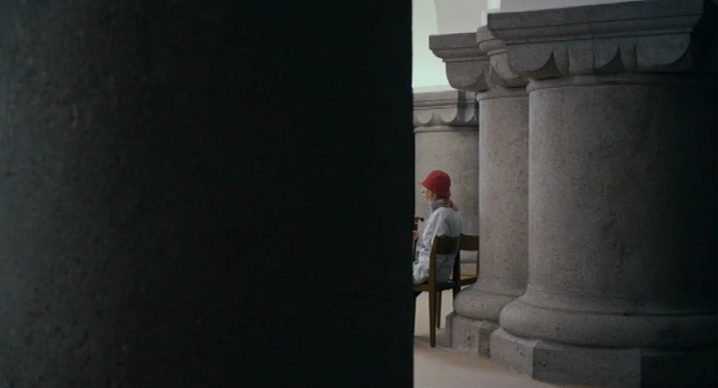 Lourdes movie still: wide — A stone colonnade features large circular pillars made of rough hewn granite blocks. A person wearing a red hat sits on…