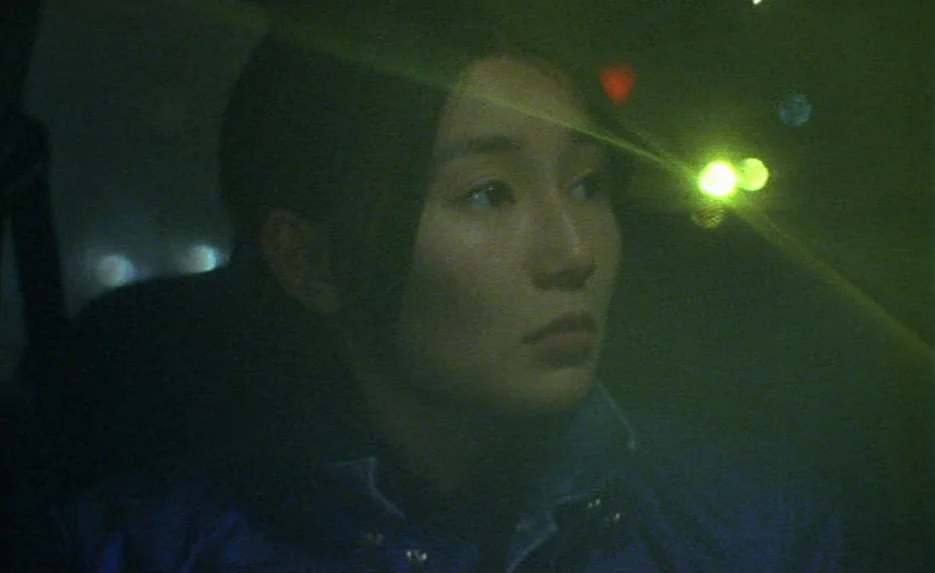 Irma Vep (1996) movie still: closeup — A woman sits inside a car at night with a green light cast over her. She looks forward while city lights…