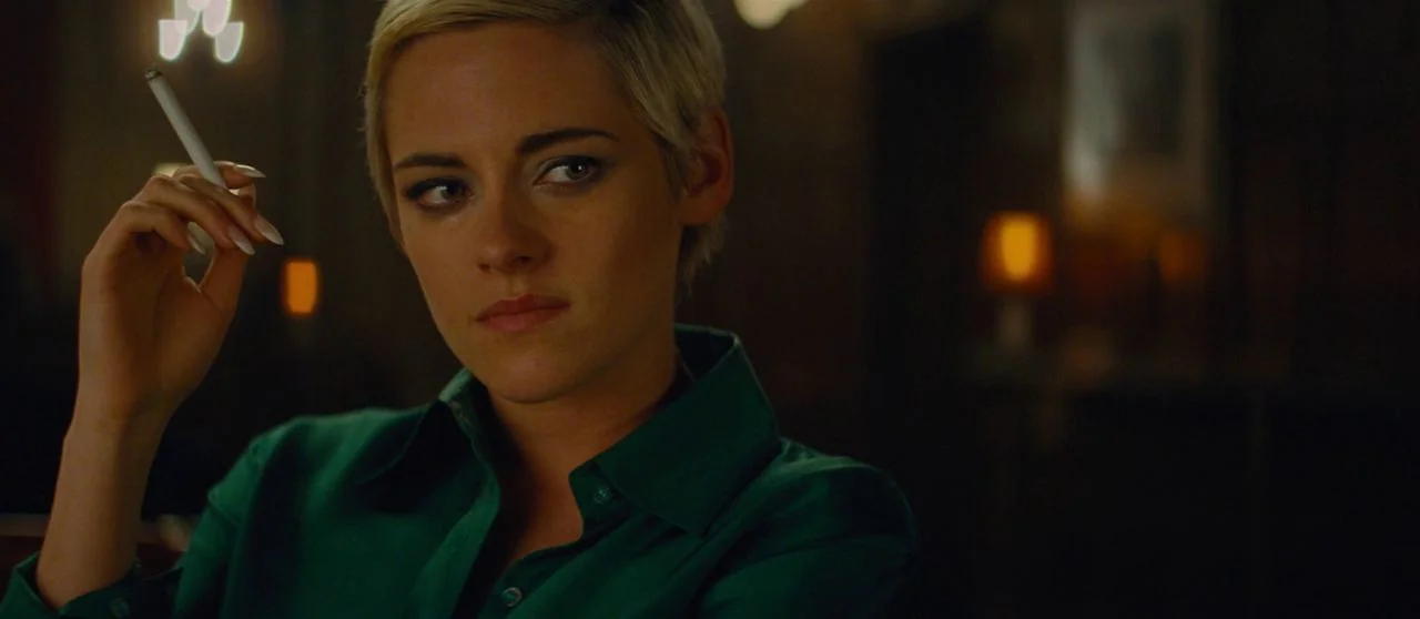 Seberg (2019) movie still: closeup — A dimly lit lounge with blurred golden light sources in the background. A woman with short blonde hair looks…