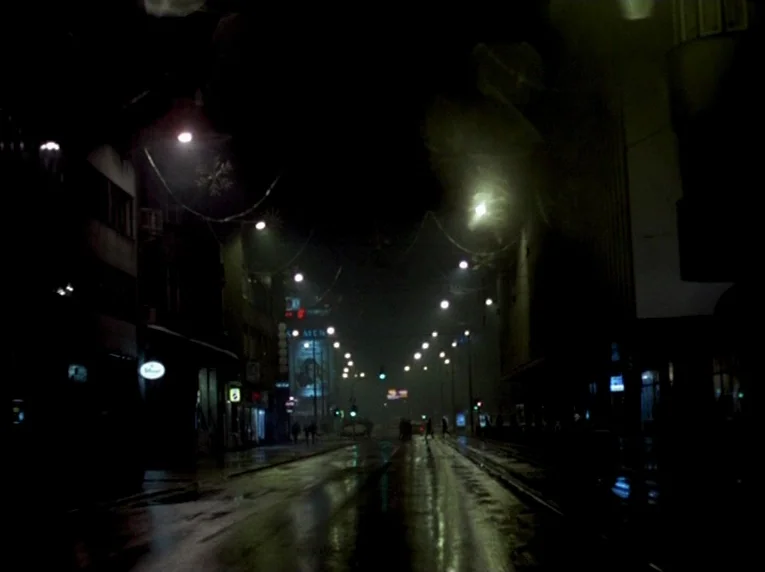 Notre Musique (2004) movie still: wide — A dark, wet city street at night is lined with buildings and overhead cables. Rows of streetlights cast…