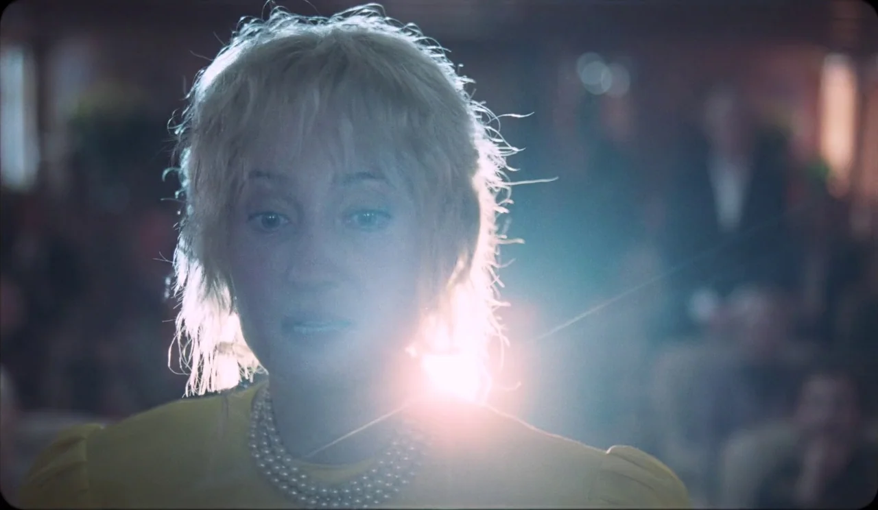 La Chimera (2023) movie still: closeup — A woman stands in a dimly lit auditorium with a blurred audience behind her. She is illuminated by intense…