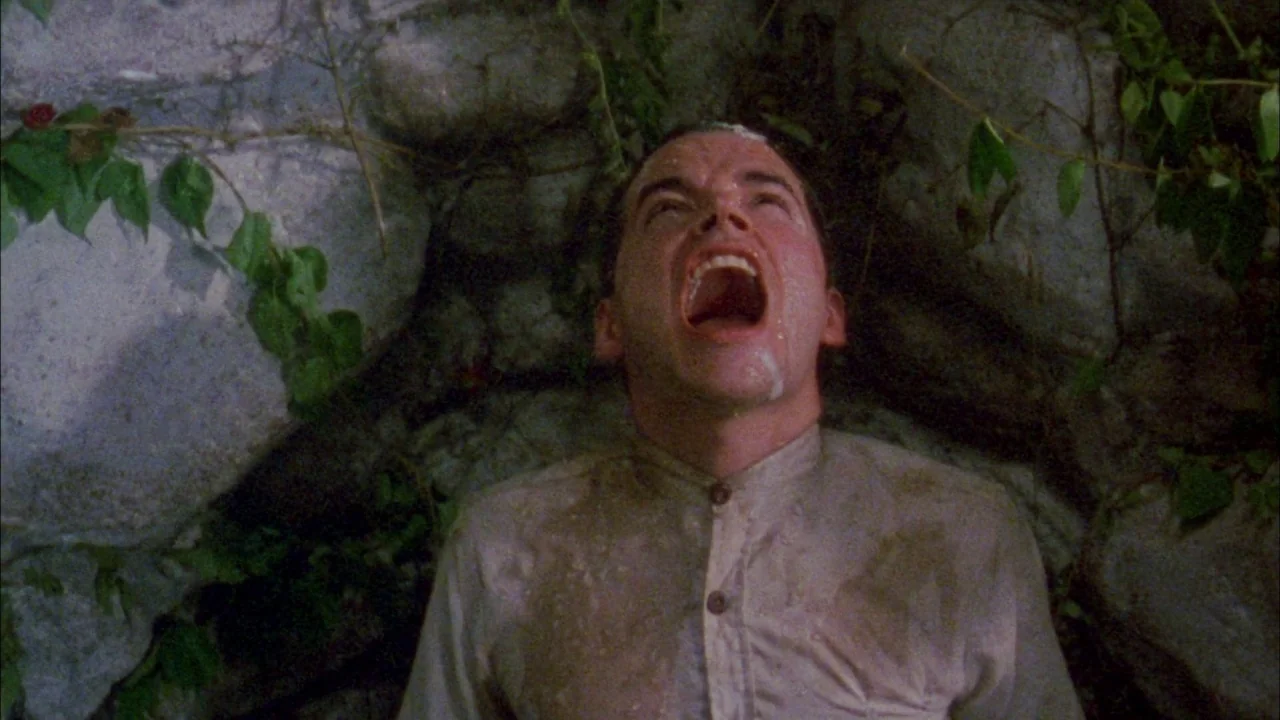Poison movie still: medium — A man stands against a rough, dark stone wall covered in climbing green vines. The subject tilts his head back with his…
