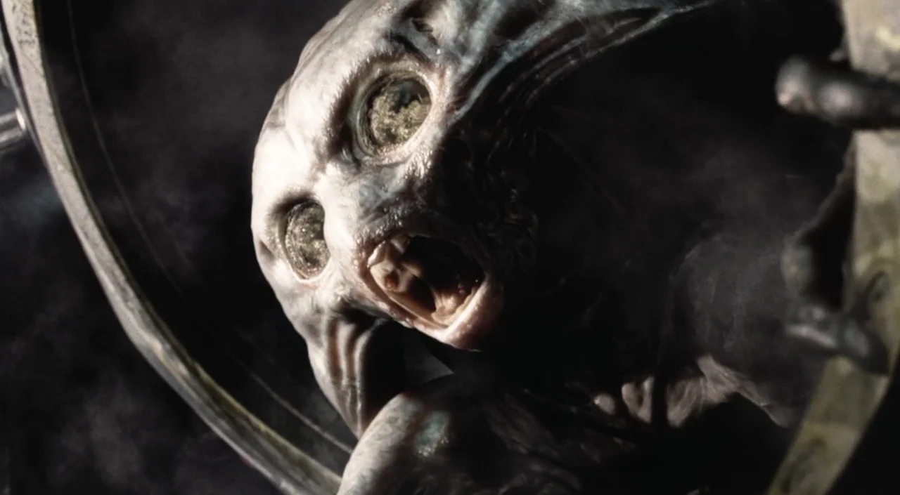 War of the Worlds (2005) movie still: closeup — A dark, cramped space surrounds a grey creature with wide eyes peering through a portal. The subject…