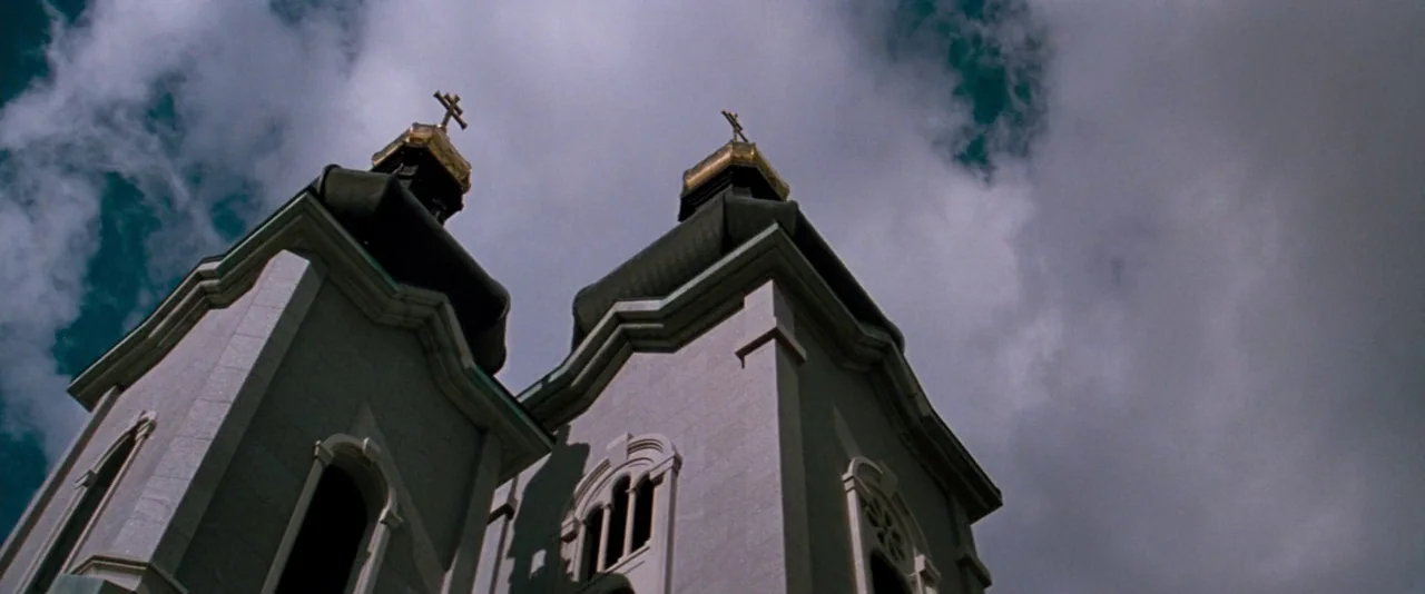 In The Mouth of Madness (1994) movie still: establishing — Two light-colored stone church towers stand against a sky filled with grey clouds.…