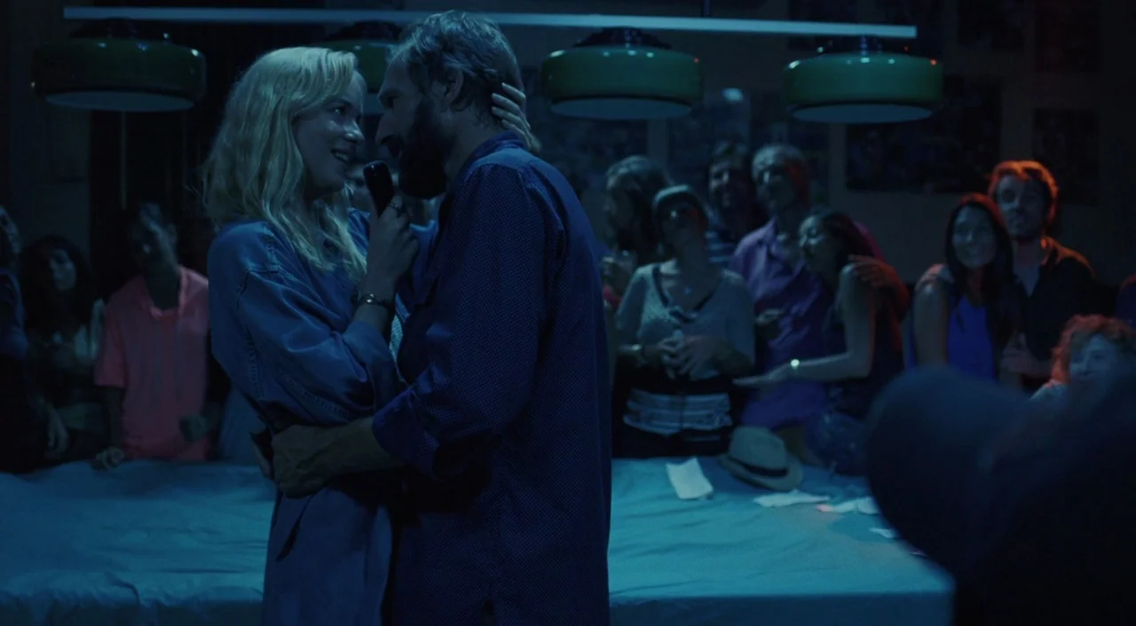 A Bigger Splash (2015) movie still: medium — A crowded bar interior with low hanging green lamps illuminates a dimly lit space. A man and a woman…