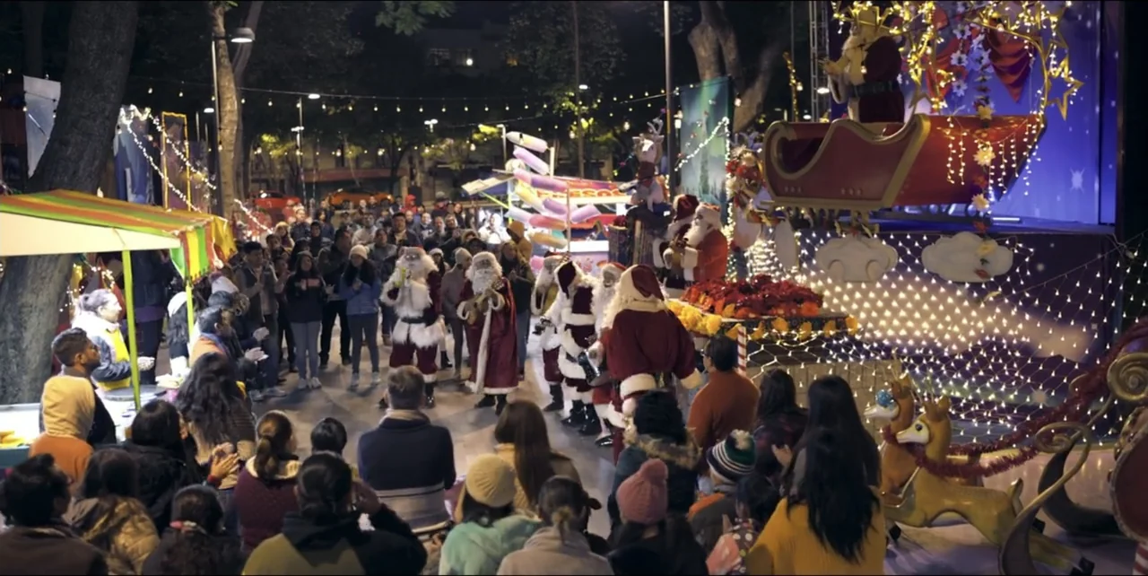 El sabor de la Navidad (2023) movie still: wide — A public plaza at night features a festive stage setup with multiple Santa performers. A large…