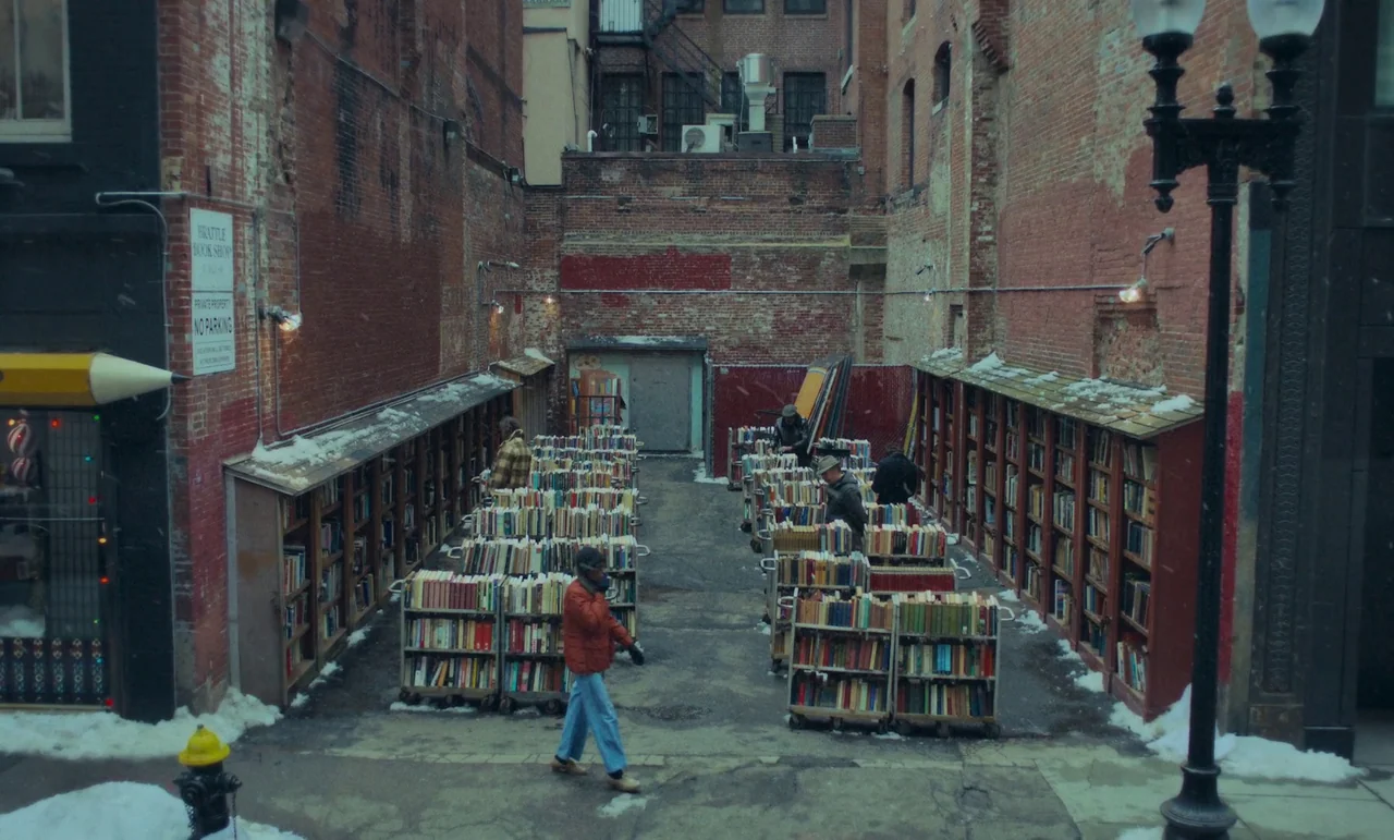 The Holdovers (2023) movie still: wide — An outdoor bookstore in a narrow urban brick alley during a light snowfall. Multiple rows of rolling book…