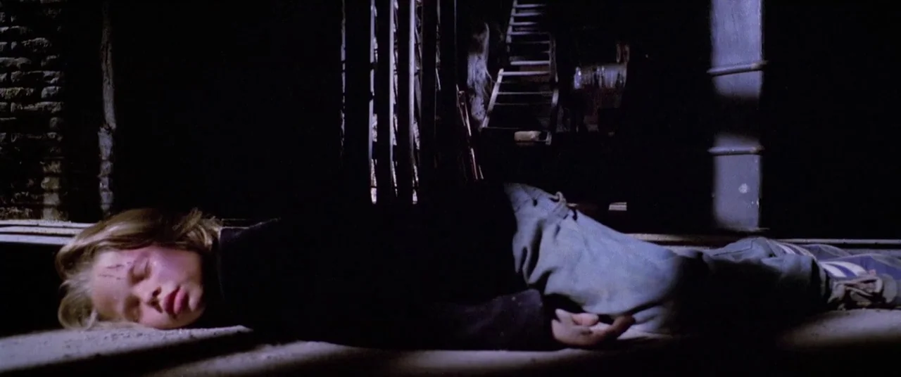 House By The Cemetery (1981) movie still: medium — A dark, unfinished room with a wooden staircase in the background. A child is lying on the floor…