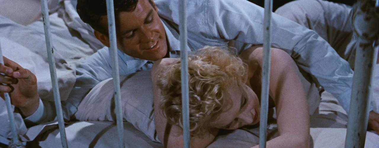 Bus Stop (1956) movie still: medium — A white metal bed frame obscures the view of a man and woman. The man is lying behind the woman, embracing her…