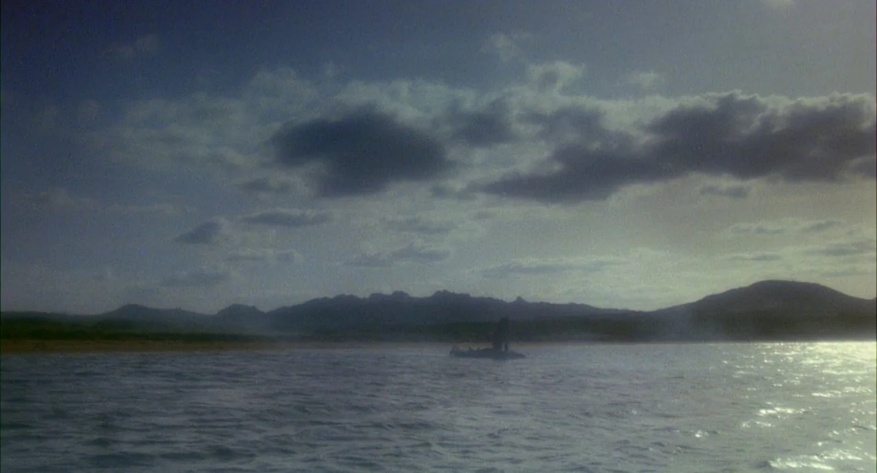Conquest (1983) movie still: establishing — A wide view shows the ocean surface with a distant shoreline and jagged mountains. A small boat is…