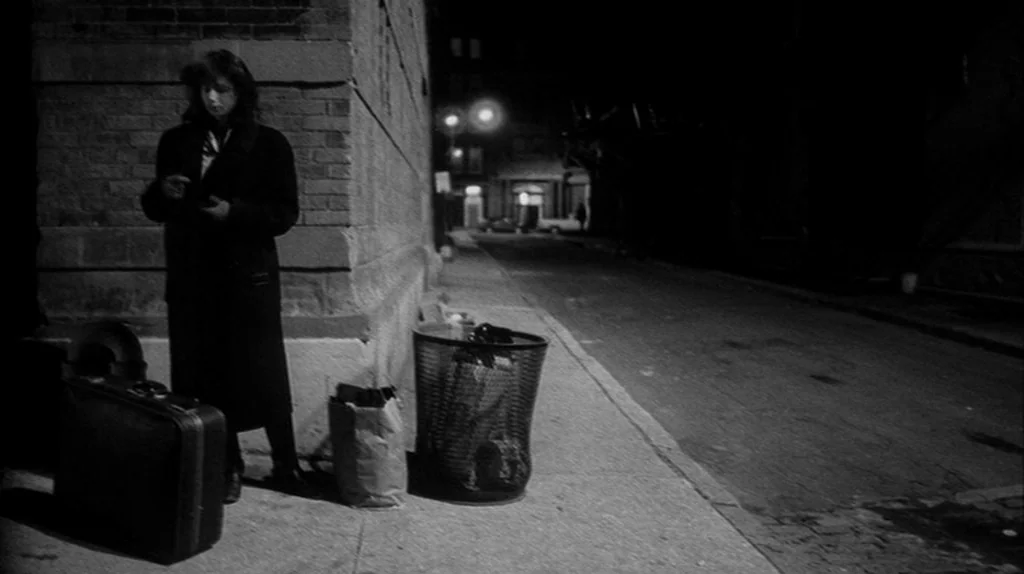 Stranger Than Paradise (1984) movie still: fullbody — A quiet urban sidewalk at night features a dark brick corner of a building. A woman stands…